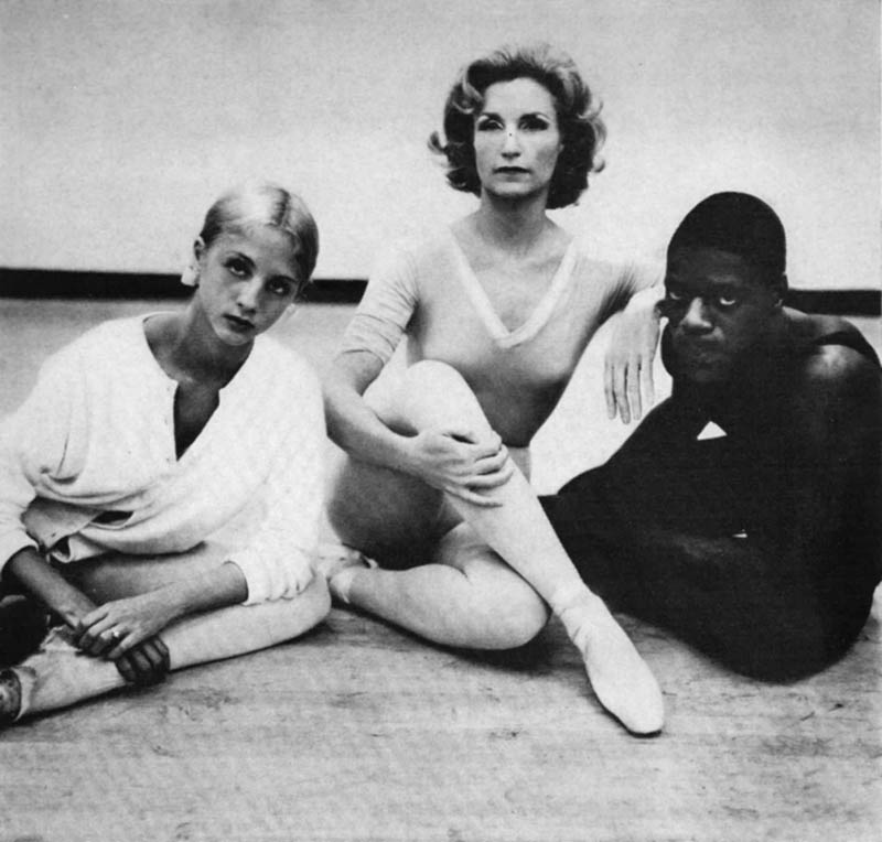 Three ballet dancers sit on the floor posed and look at the camera.