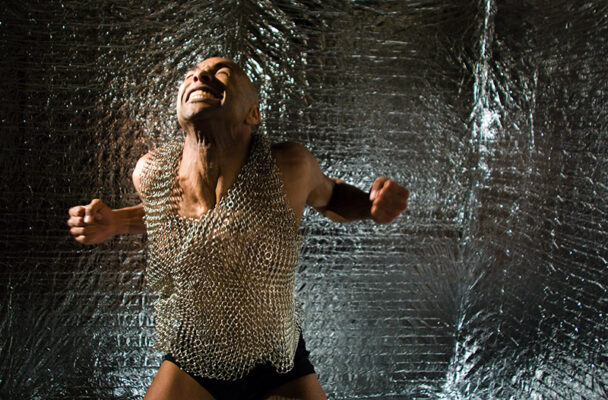 Darrell wears a chainmail shirt and black shorts and looks as if he is breaking free of the shirt, his body flexed and his wrists in fists.