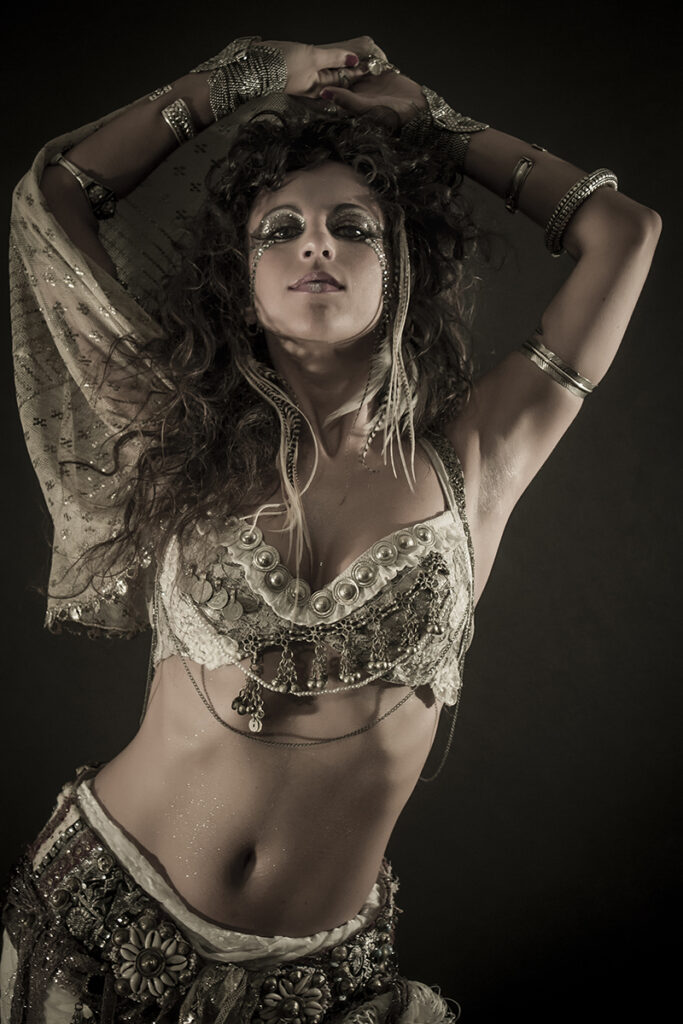 Alhazar poses with her arms above her head and wearing a gold belly dance costume.