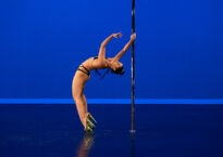 Irlanda does a backbend on a pole. She is wearing a black bikini and gold Pleaser heels, and the background is blue.