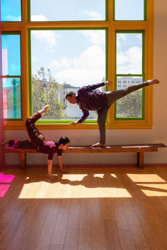 Two dancers on a bench by a huge window. One is standing in a penchee arabesque reaching for the other, who is in a push up position with one leg extended above.
