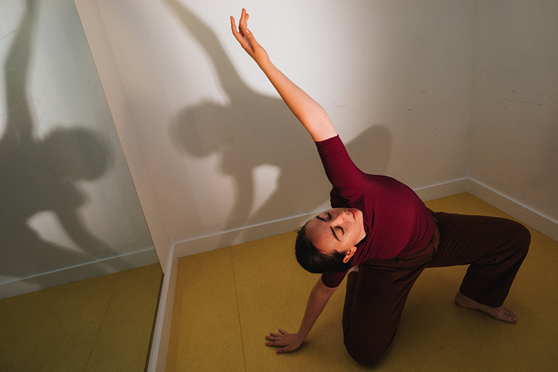 A dancer crouches in a kneel, one arm and face extended upward. Their shadow is cast on the wall behind them.