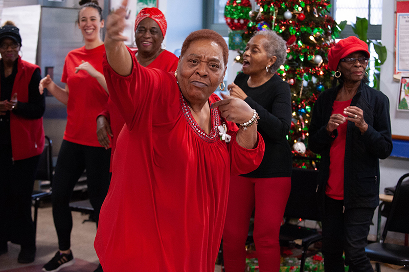 An older dancer in red gestures forward with charisma. A Christmas tree is in the background.