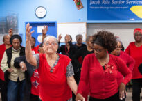 Two older dancers wearing red lead a group of people behind them in a senior center.
