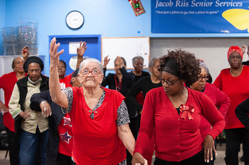 Two older dancers wearing red lead a group of people behind them in a senior center.