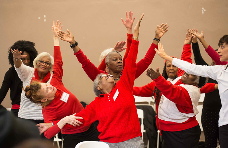 A group of older dancers wearing red lift their arms in various directions and look upward.
