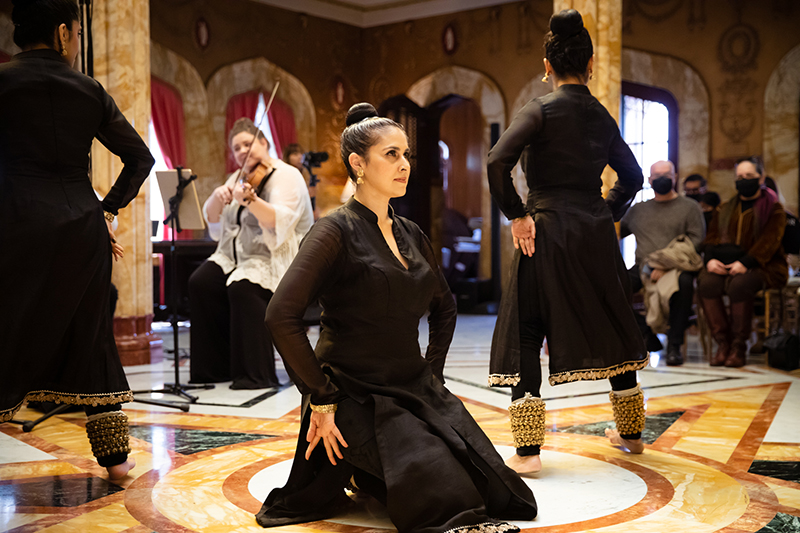 Rukhmani kneels in the center of several dancers and musicians. She wears a stiff black dress.