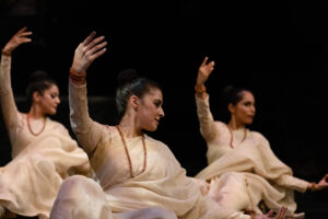 Three dancers wearing white dresses lift one arm and look over their shoulders.