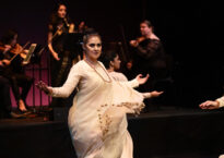 Rukhmani dances with open arms in front of musicians. She wears a white flowing dress.