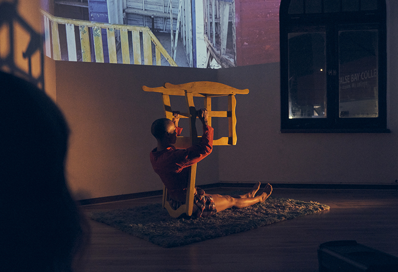 Lorin is seated on the ground holding a chair upside down above her. A projection is in the background.