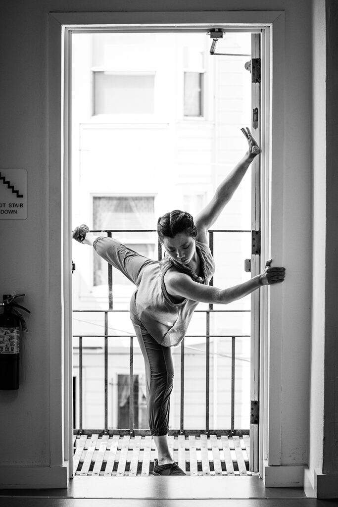 A black and white photo of a dancer in a doorway with two hands on the sill and her leg behind her out the door.