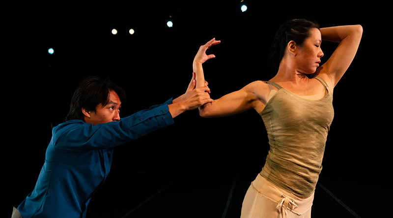 One dancer stands and leans away with both arms elevated while another dancer pulls on an arm providing counterbalance.