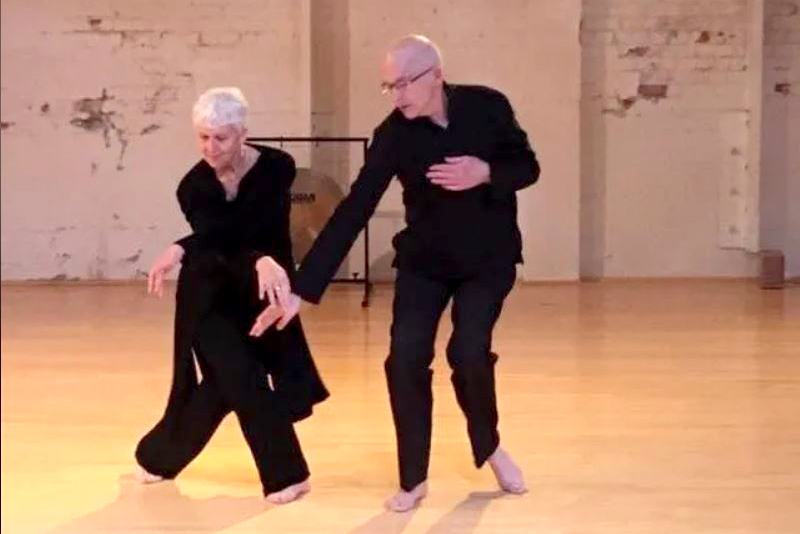 A man and a woman on a wood floor dancing. Both are wearing black and reaching hands downward toward each other.