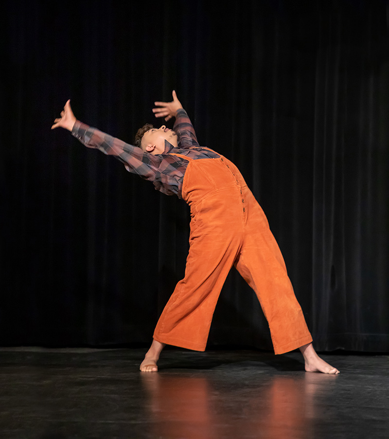 A dancer wearing overalls stands in a wide stance on stage and reaches up and behind them.
