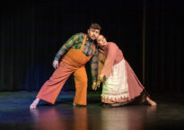 Two dancers wearing 19th century costumes lean into each other onstage.