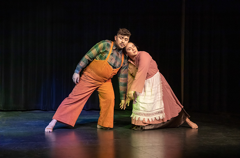 Two dancers lean into each other onstage. One is wearing overalls and the other a dress and apron.