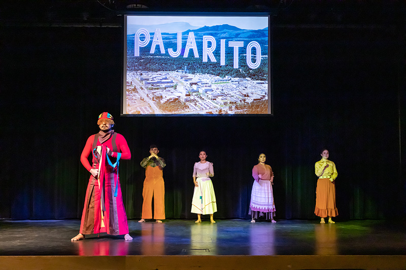 Five dancers stand in wide stances facing forward onstage wearing an assortment of colorful costumes. A projection with the words "Pajarito" is above them.