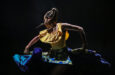 A dancer in a yellow shirt and blue skirt whips her braids to the side under dramatic lighting.