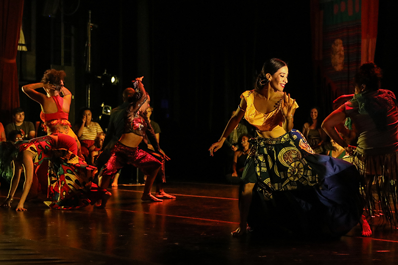 A low lit stage with dancers moving in vairous colorful patterned outfits.