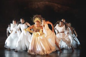 Several dancers wearing white and in a triangle formation lunge forward.