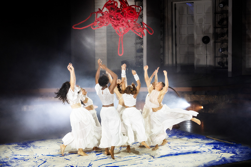 Several dancers wearing white jump up, reaching toward a red dangling mess of rope.
