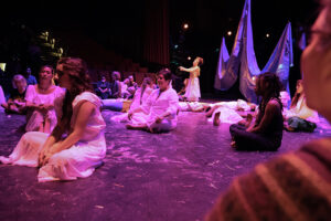 Many people sit onstage facing various directions, many are wearing white.