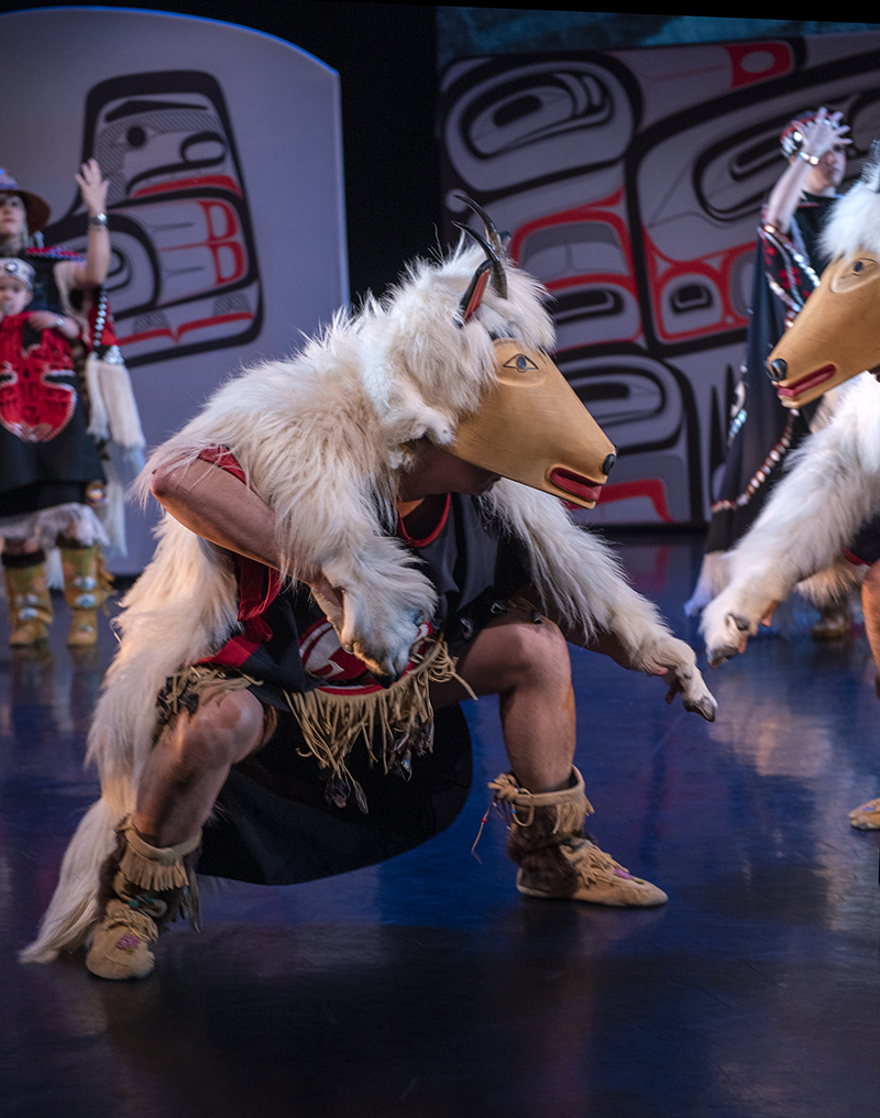 A dancer wearing animal regalia squats low on the stage, facing another person in animal regalia barely in the frame.