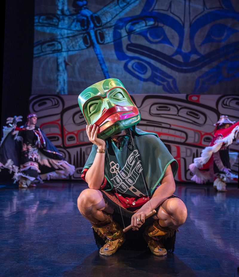 A dancer wearing a green mask and regalia crouches on the stage, with other dancers moving behind.