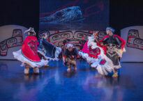 Dancers wearing red and white regalia dance in front of a projection of a beaver.