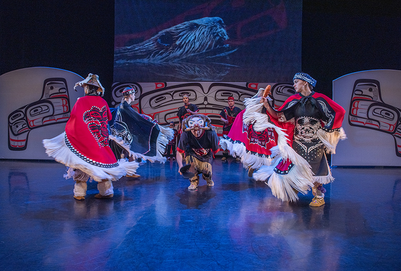Dancers wearing red and white regalia dance in front of a projection of a beaver.