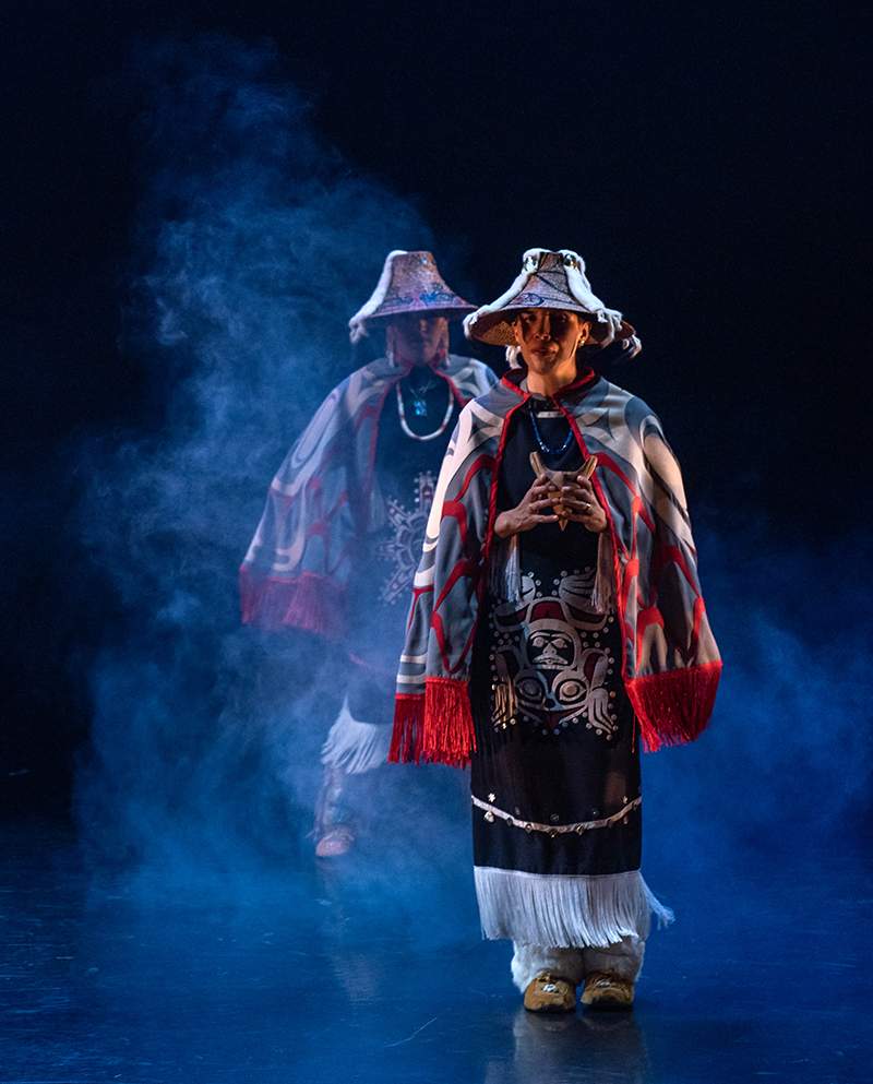 A dancer stands in regalia in a mist on stage, with another dancer standing in the background.