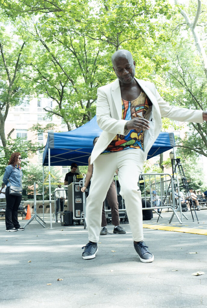 Sekou dances in a white outfit on an outdoor stage with trees in the background.