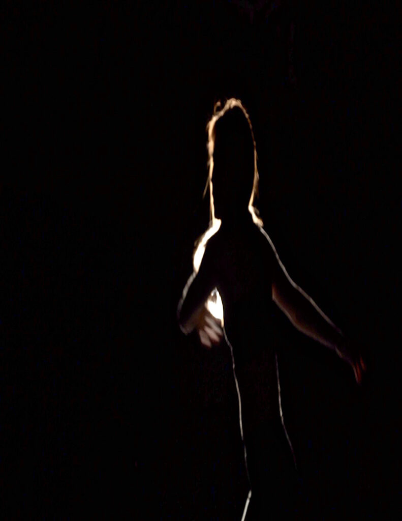 A backlit blurry image of a dancer with arms as if she's about to throw herself into movement.