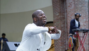 Sekou smiles while performing in a white jacket. He brings his arms up under his chin.
