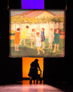 Image Description: Vanessa and her walker Pluto are silhouetted as they travel away from the viewer and through a narrow portal of fiery sunlight at the back of the stage, under a large tapestry print of Petrona Viera’s Recreo (c. 1924) displaying a hopeful vision of children dancing through and around each other in the lush green grass.