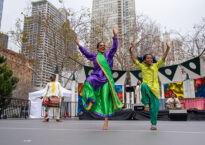 A drummer in the background with two brightly dressed dancers in the foreground with one leg raised and both arms raised. They perform outside with skyscrapers in the background.
