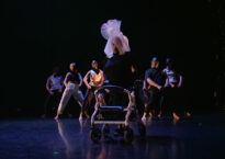 Image Description: A photo of Vanessa on stage with her walker named Pluto. She is wearing all black and has a white mesh head piece that looks like an abstract Venus fly trap. Her walker has a costume on that is a white fabric looped around the purple metal parts of her walker. In the background, there are six dancers in a wide stance leaning back.