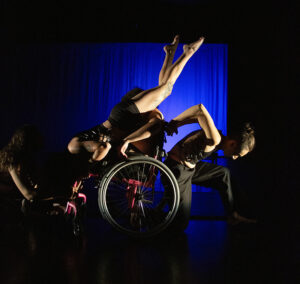 Image Description: Orli supports Zera, who is hanging upside down from Orli's wheelchair, legs up and dangling in the air. Martin and Ronan tilt Orli on opposite sides. Behind them is a dark blue backdrop.