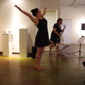 Lauren stands on tiptoe with arms in a V above her head. Clara plays bass clarinet in the backfround. They both wear black on a hard wood floor.