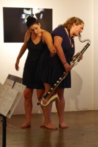 Lauren stands close to a bass clarinet player who is also standing. Lauren gestures forward. They wear black and are on a wood floor.