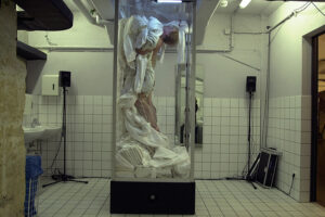 Two dancers pose in a vertical glass rectangle in a public bathroom.