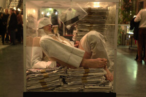 A dancer in a glass cube filled with newspaper is scrunched down against the glass. A busy metrolopitan