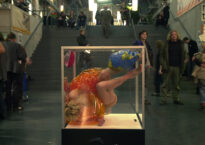 A woman in a glass cube moves almost upside down. Around her, people walk up and down public stairs.