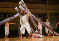 Dancers wearing white move around the stage lifting long tube-like structures.