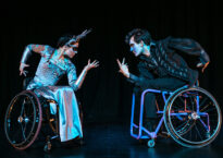Two dancers in wheelchairs face off on a black stage. One is wearing a silver dress and headdress, the other is wearing black with face makeup.