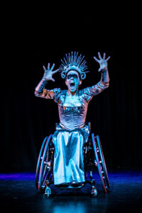 A dancer in a wheelchair and a silver dress dramatically raises her hands like claws at the audience.