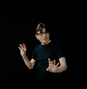 A man wearing black and glasses with claw-like fingers on his arms extended a little in front of him. He is against a black backdrop.