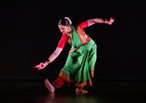 Sloka bends over one leg, gesturing toward her flexed foot. She wears a green and red sari.