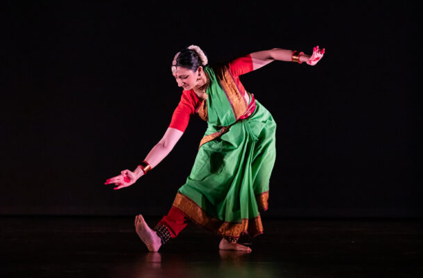 Sloka bends over one leg, gesturing toward her flexed foot. She wears a green and red sari.