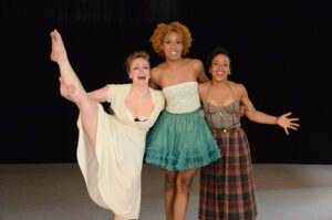 Three dancers pose and smile on stage, one does a leg lift.